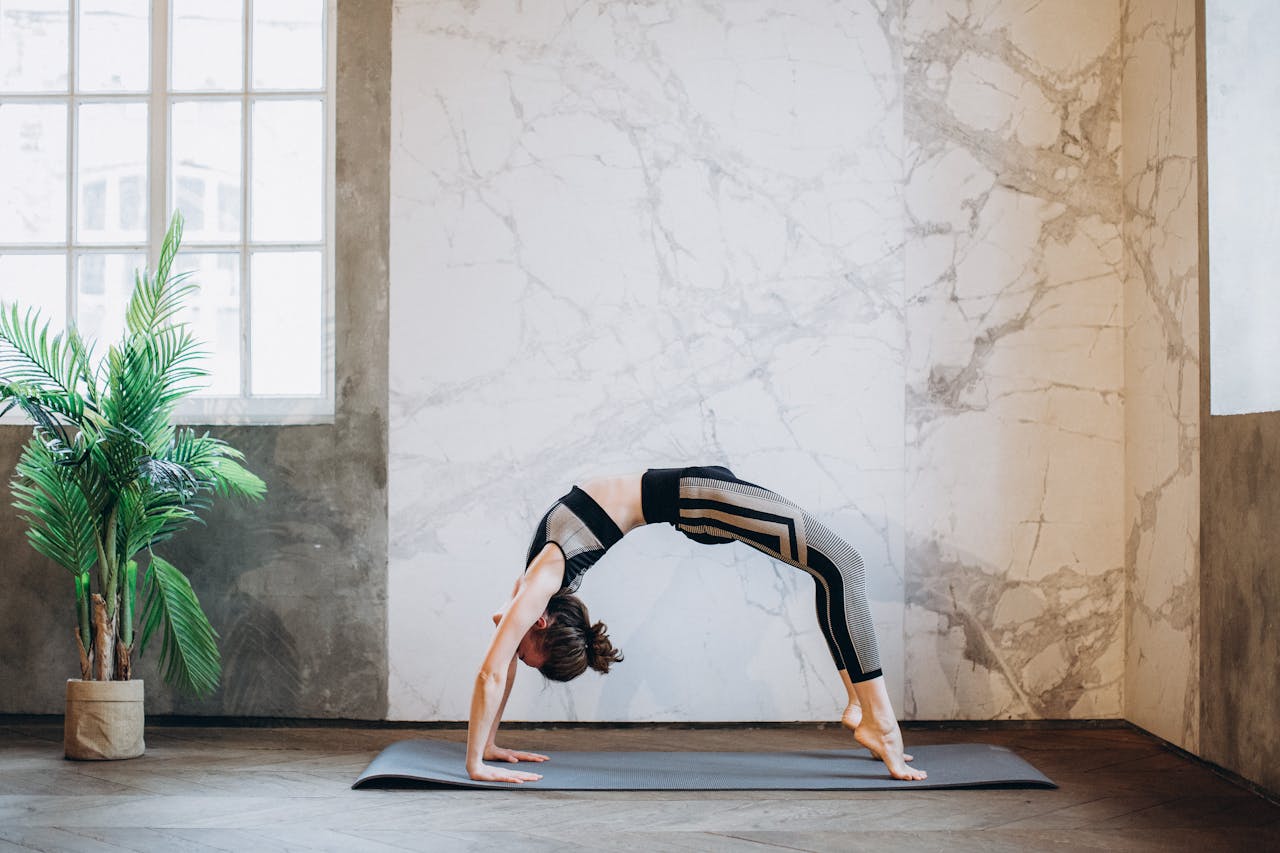Adult woman practicing yoga backbend pose in a serene indoor space with a potted plant and large window.